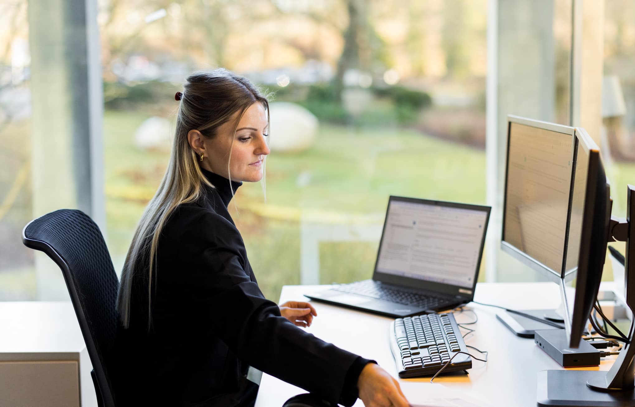 Een vrouwelijke collega van Van Havermaet zit achter haar bureau in een moderne kantoorsetting. Ze draagt een zwarte coltrui en heeft blonde haren. Voor haar staan twee schermen en een laptop.